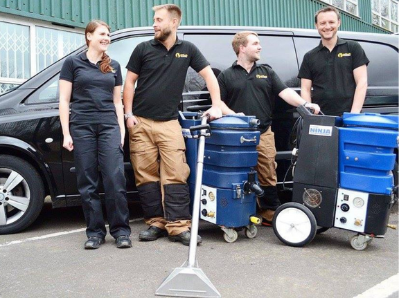 This is a photo of Clerkenwell Carpet Cleaning carpet cleaners (three men and one woman) standing in fromt of their black van, with two steam cleaning carpet machines next to them